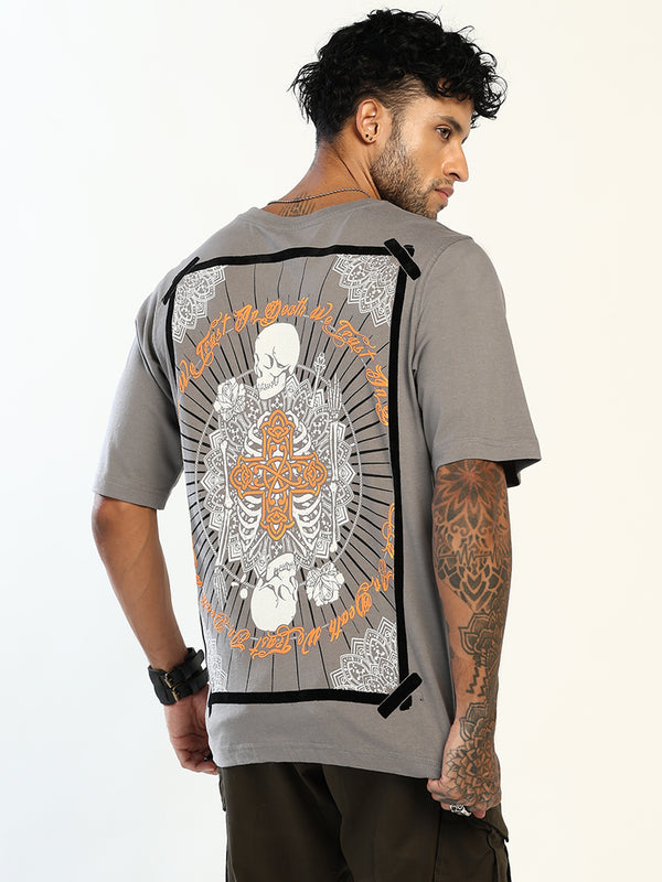 A man wearing a gray t-shirt with a large back graphic, photographed from behind. The graphic features a white skeleton design with an orange cross and gothic lettering, bordered by black tape-like details.