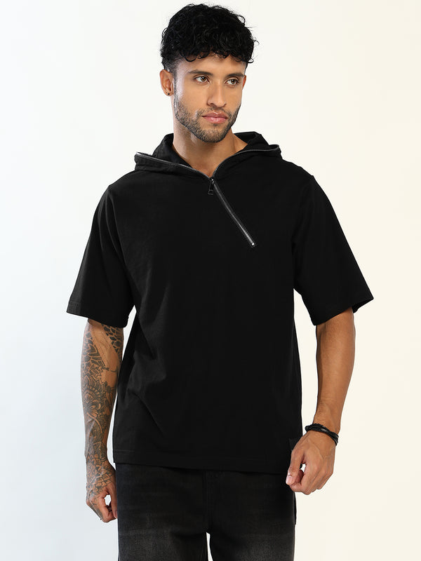 A man wearing a black short-sleeve half-zip hoodie with a silver zipper, photographed against a plain off-white studio background.