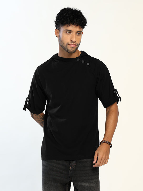 A man wearing a black short-sleeve t-shirt with unique button details on the shoulder and tie details on the sleeve cuffs, paired with dark gray jeans, against a plain white background.
